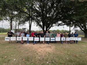 Group of people holding signs outdoors, standing in front of a tree on a grassy field.