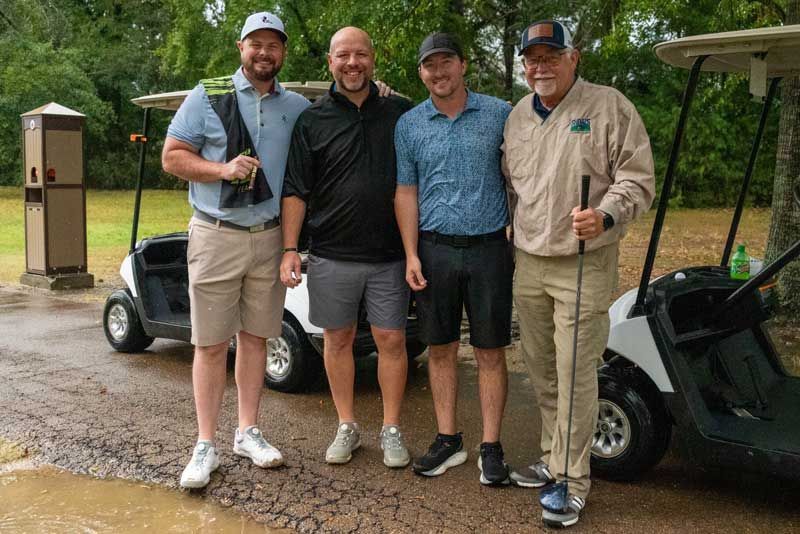 Four men stand near golf carts on a wet course. They smile, posing after golf.