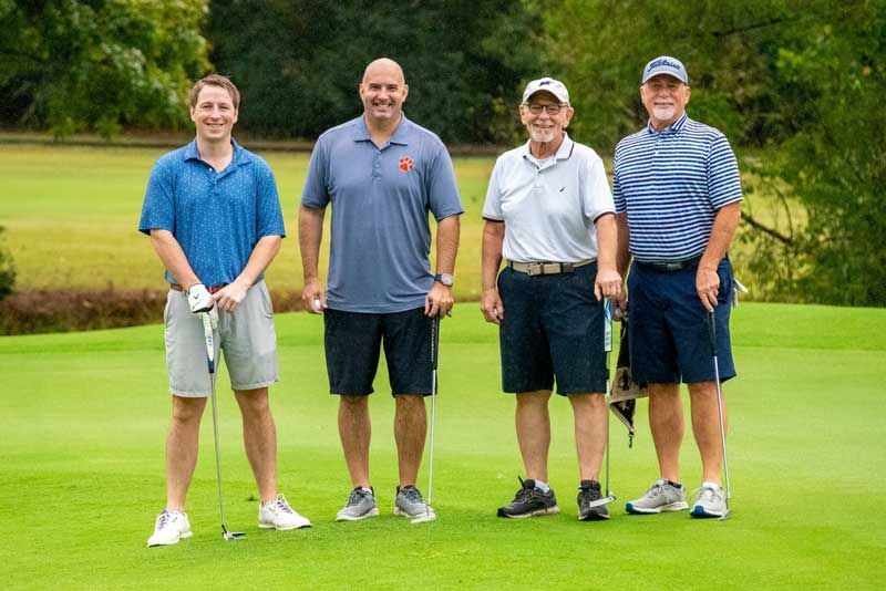 Four men in golf attire stand on a green, holding clubs.