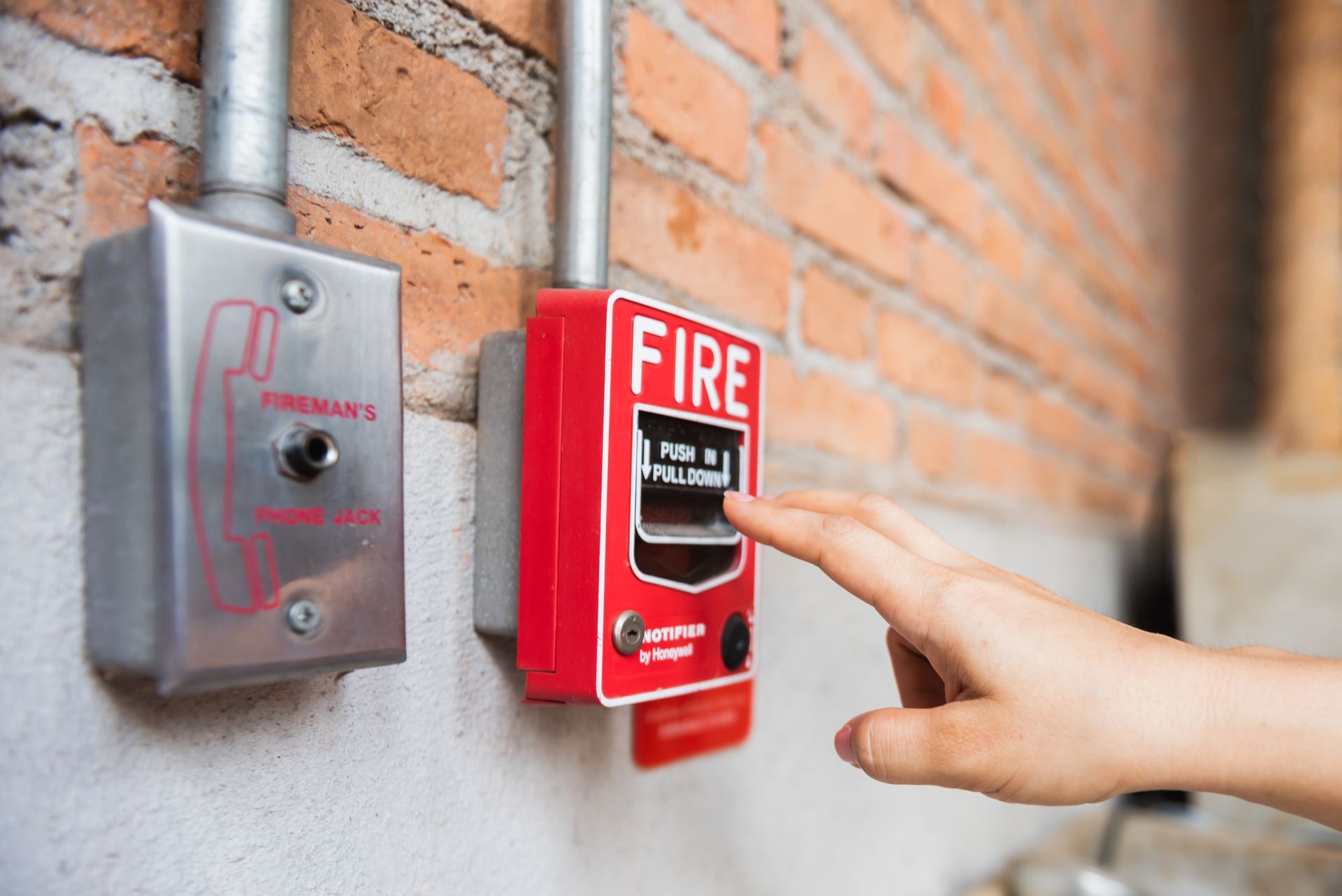 A person's hand pressing a red fire alarm button mounted on a brick wall.