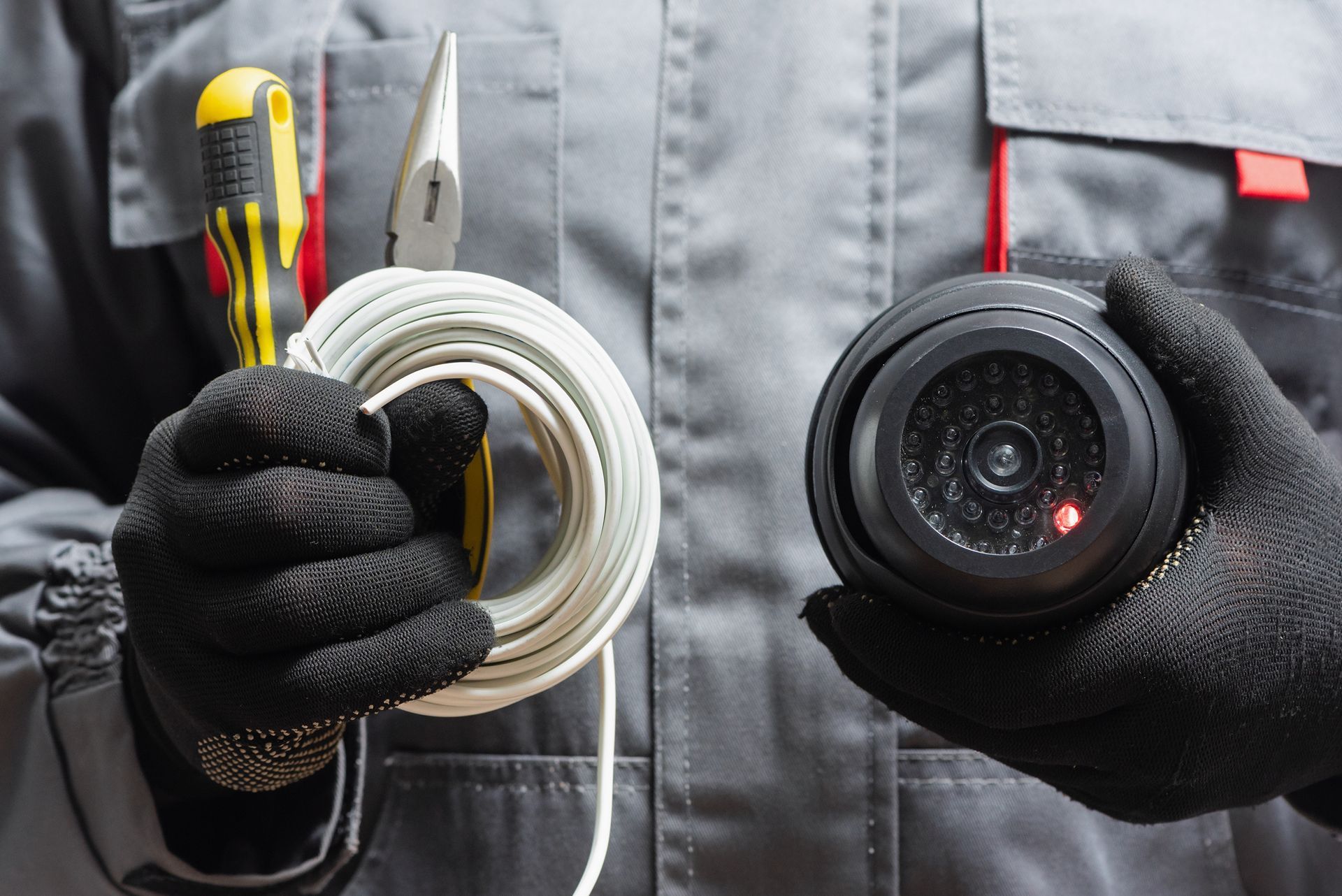 Person in work clothes, black gloves, holding security camera, wire and pliers.