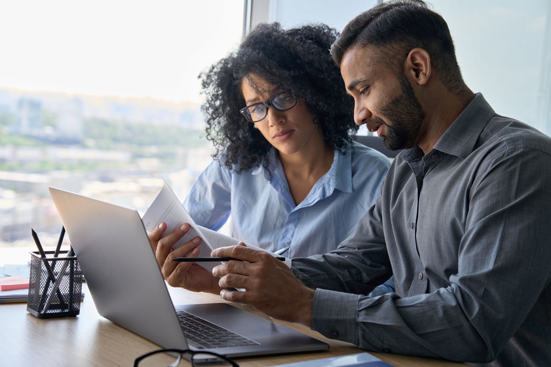 Two people looking at laptop and documents; indoor office setting.