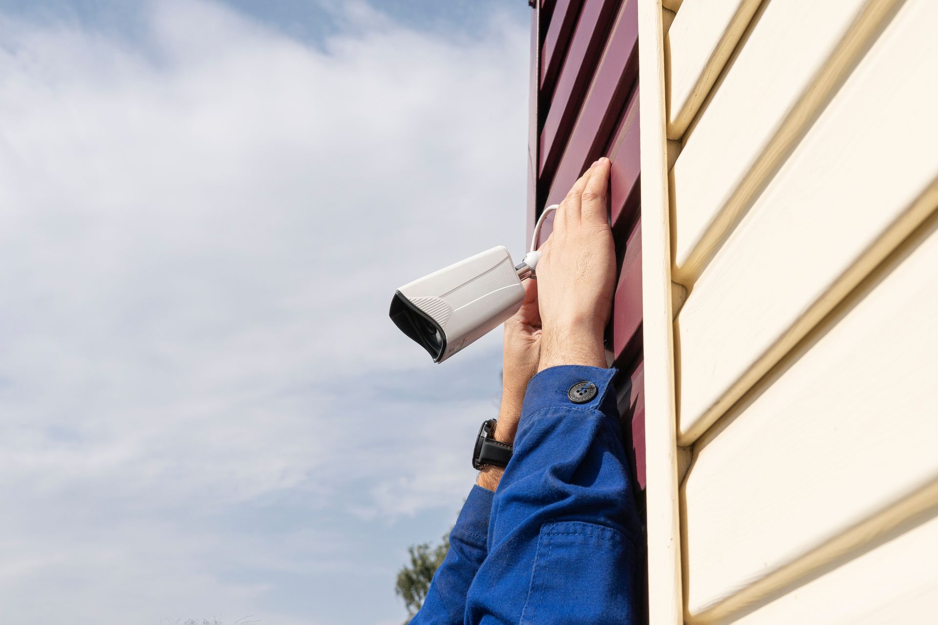 Person installing a white security camera on a tan and burgundy building.