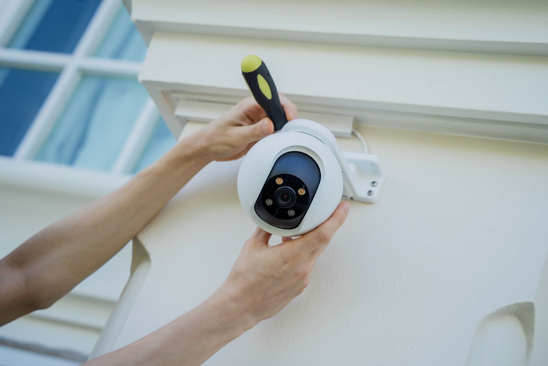 Person installing a security camera on a white building exterior with a screwdriver.