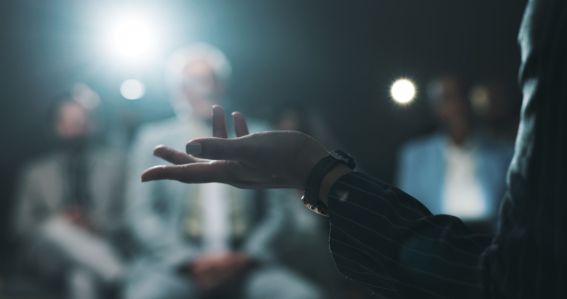 A person gestures with an open hand towards a blurred audience, lit by a spotlight.