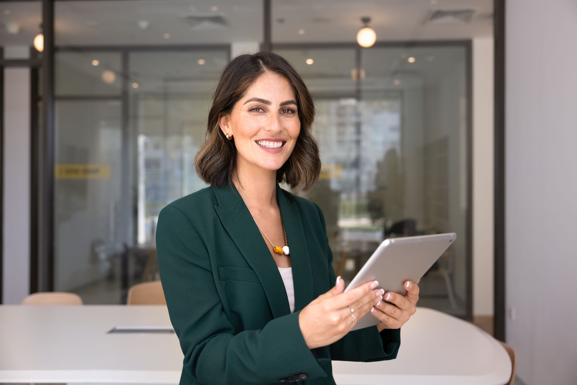 Woman in green blazer smiles, holding tablet in modern office setting.