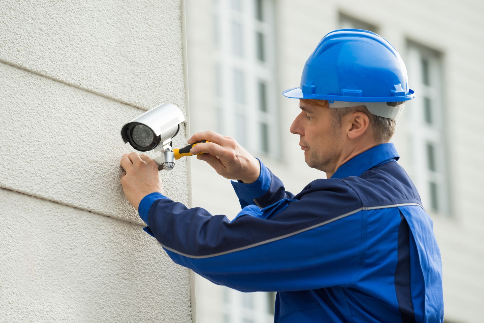Man in blue uniform and hard hat installing a security camera on a white building exterior.