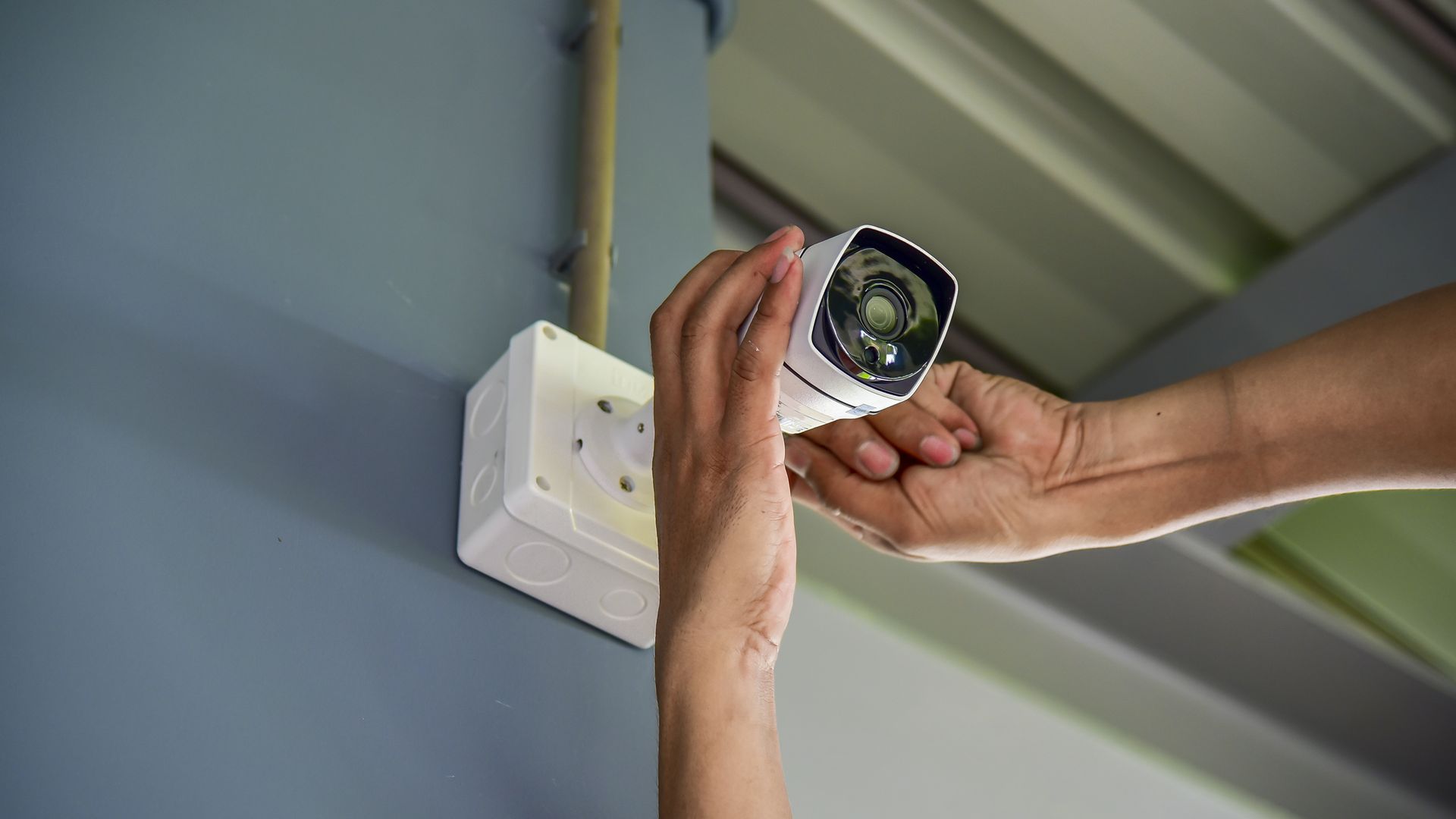 Hands mounting a white security camera on a blue wall next to an electrical outlet.