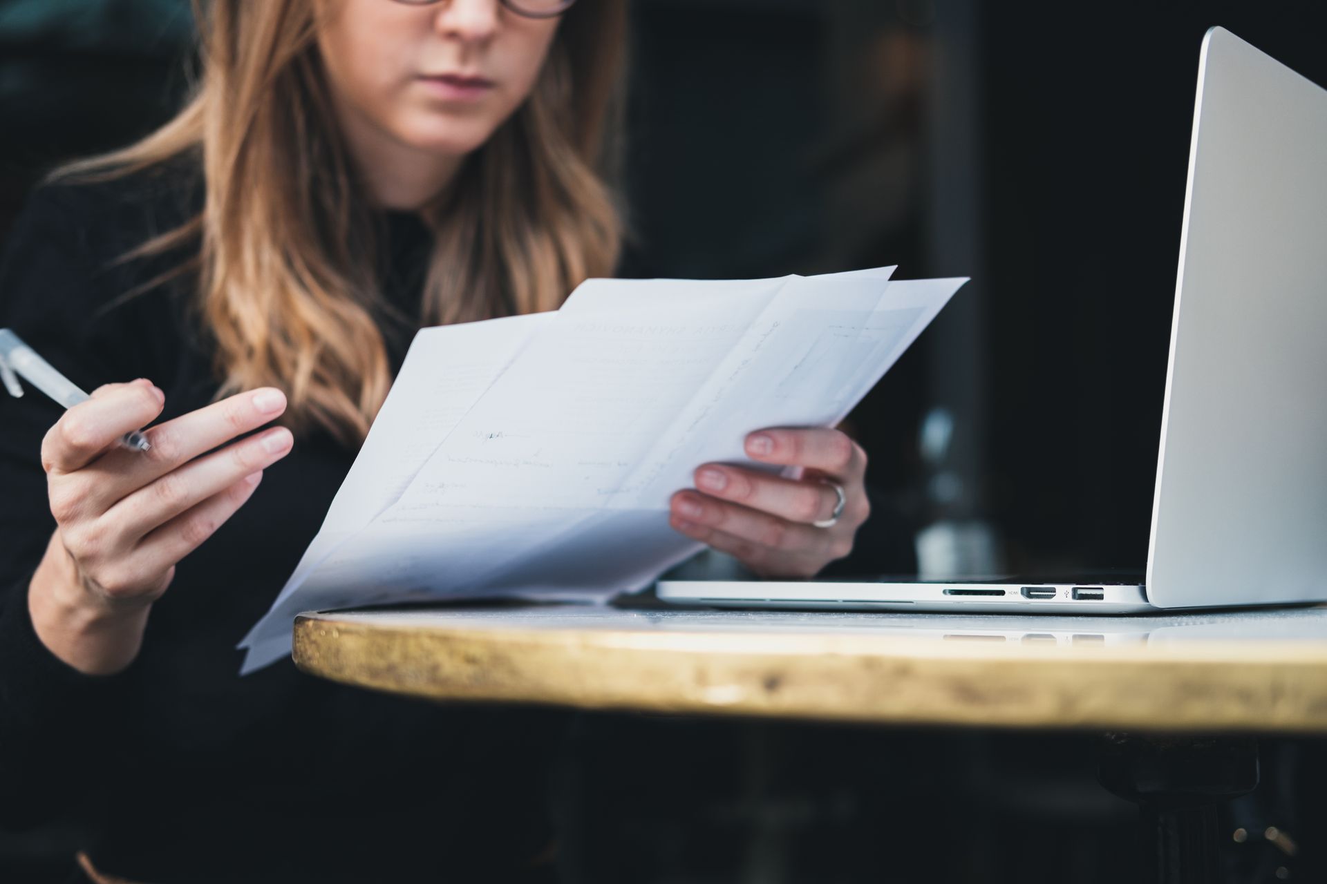 Woman with glasses reviews papers, laptop open on a table.