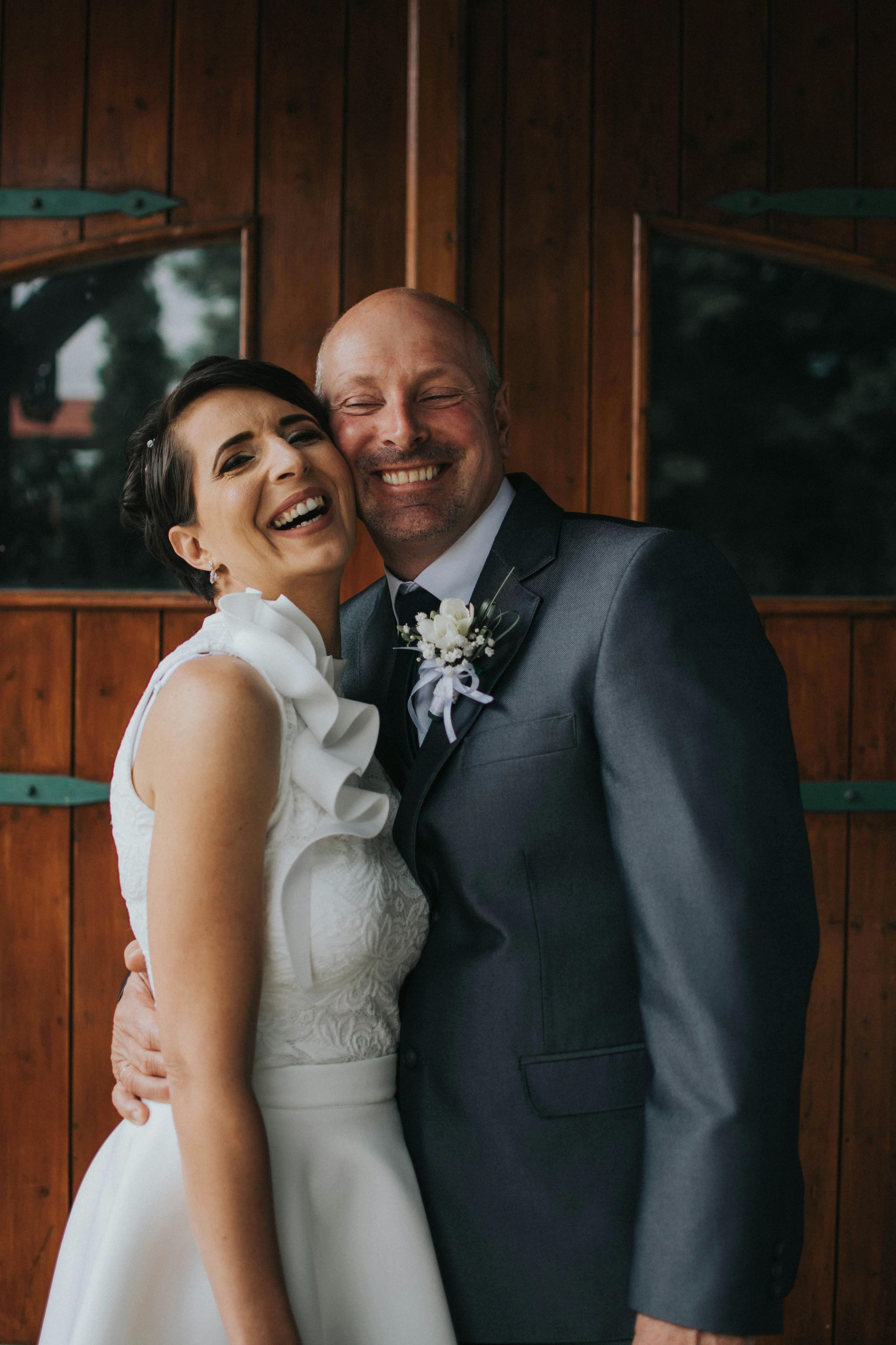Bride and groom laughing, embracing in front of wooden doors. The bride wears a white dress, the groom a grey suit.