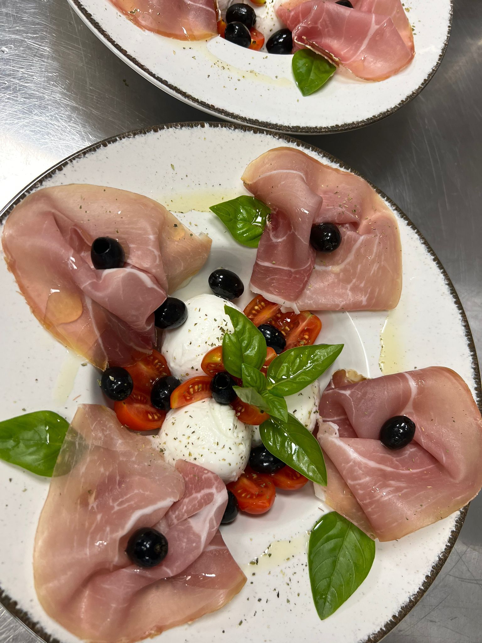 Piatti di antipasti a base di prosciutto con mozzarella, olive, basilico e pomodorini su un tavolo bianco.