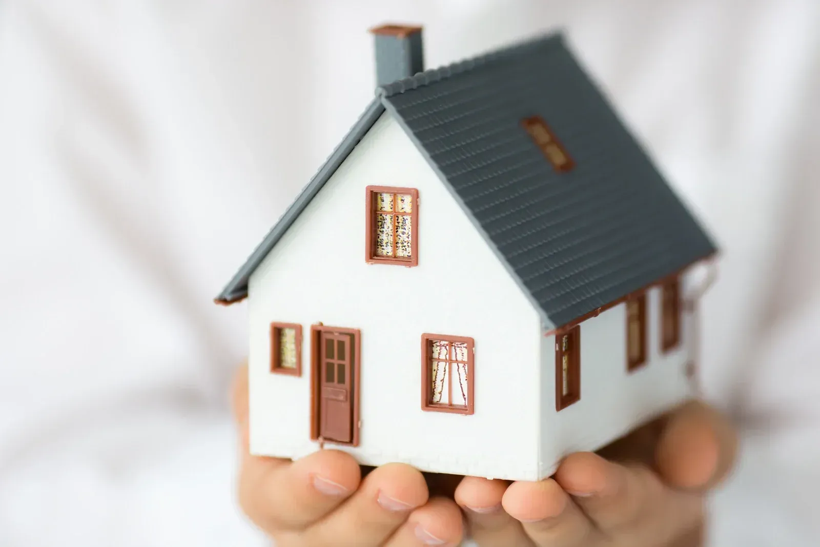 A person’s hands gently cradle a small, white model house with a grey roof against a white background.