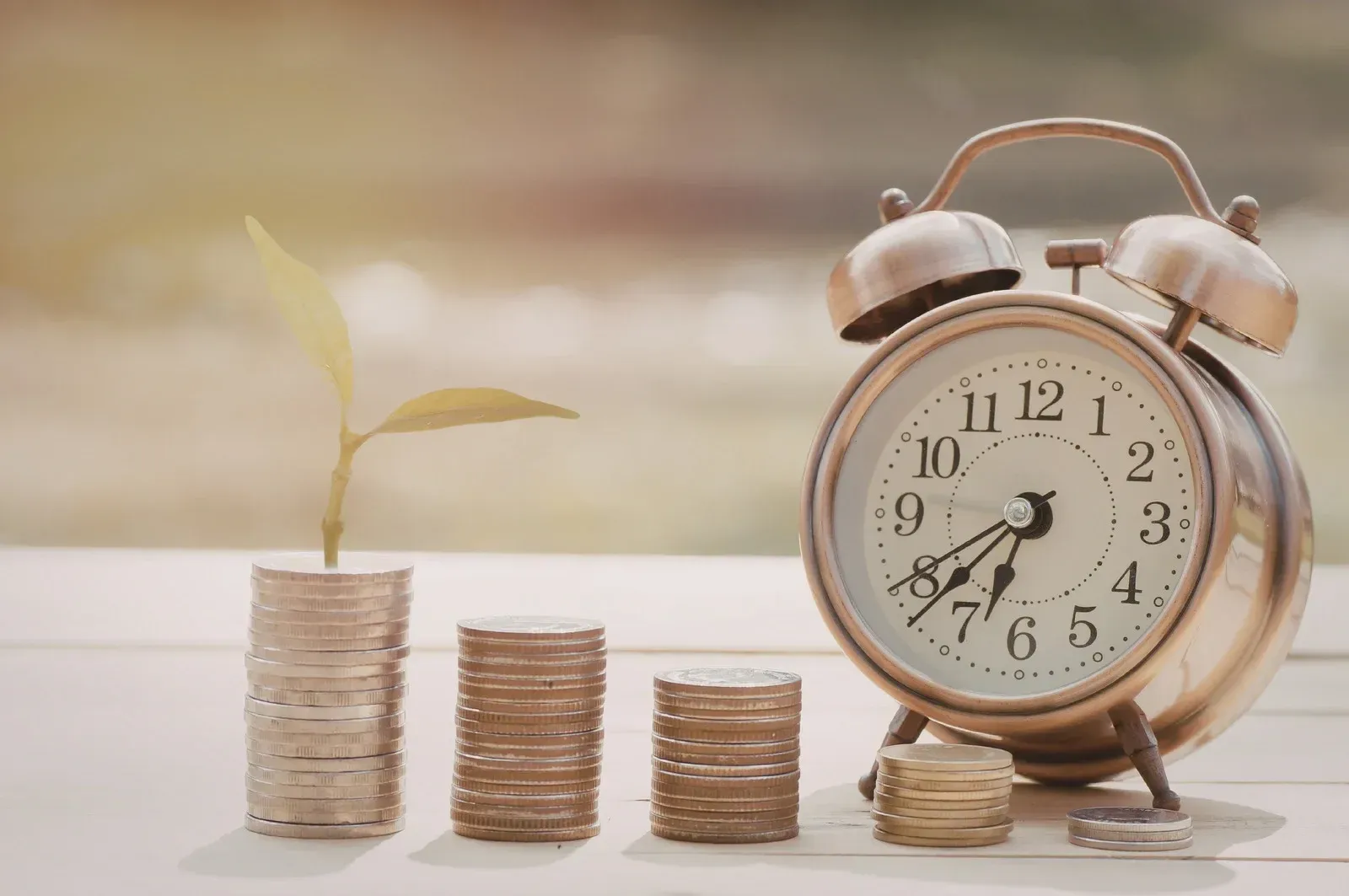 A small plant grows out of a stack of coins next to an analog alarm clock on a table, symbolizing time and investment.