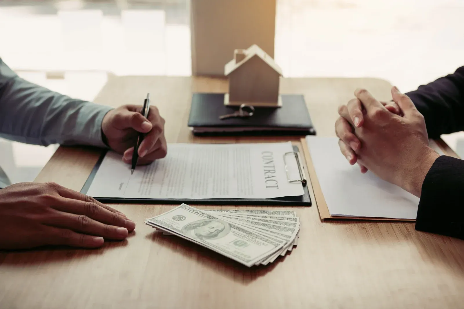 Two people sign a real estate contract at a wooden table with a miniature house model and a stack of cash.