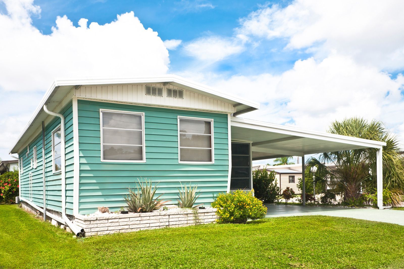 A teal mobile home with a white roof and carport, set on a grassy lawn under a bright blue sky with white clouds.