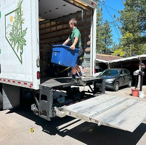 A mover carrying a blue bin down a truck ramp. The truck has a logo, a car, and a house are in the background.