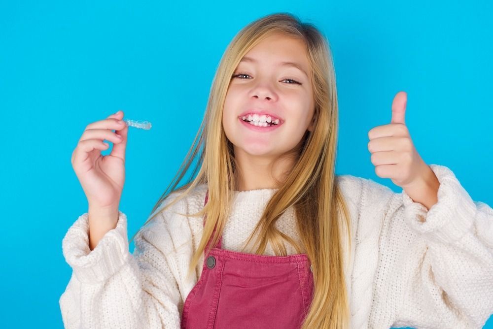 Smiling child in a pink apron gives a thumbs-up against a blue background.