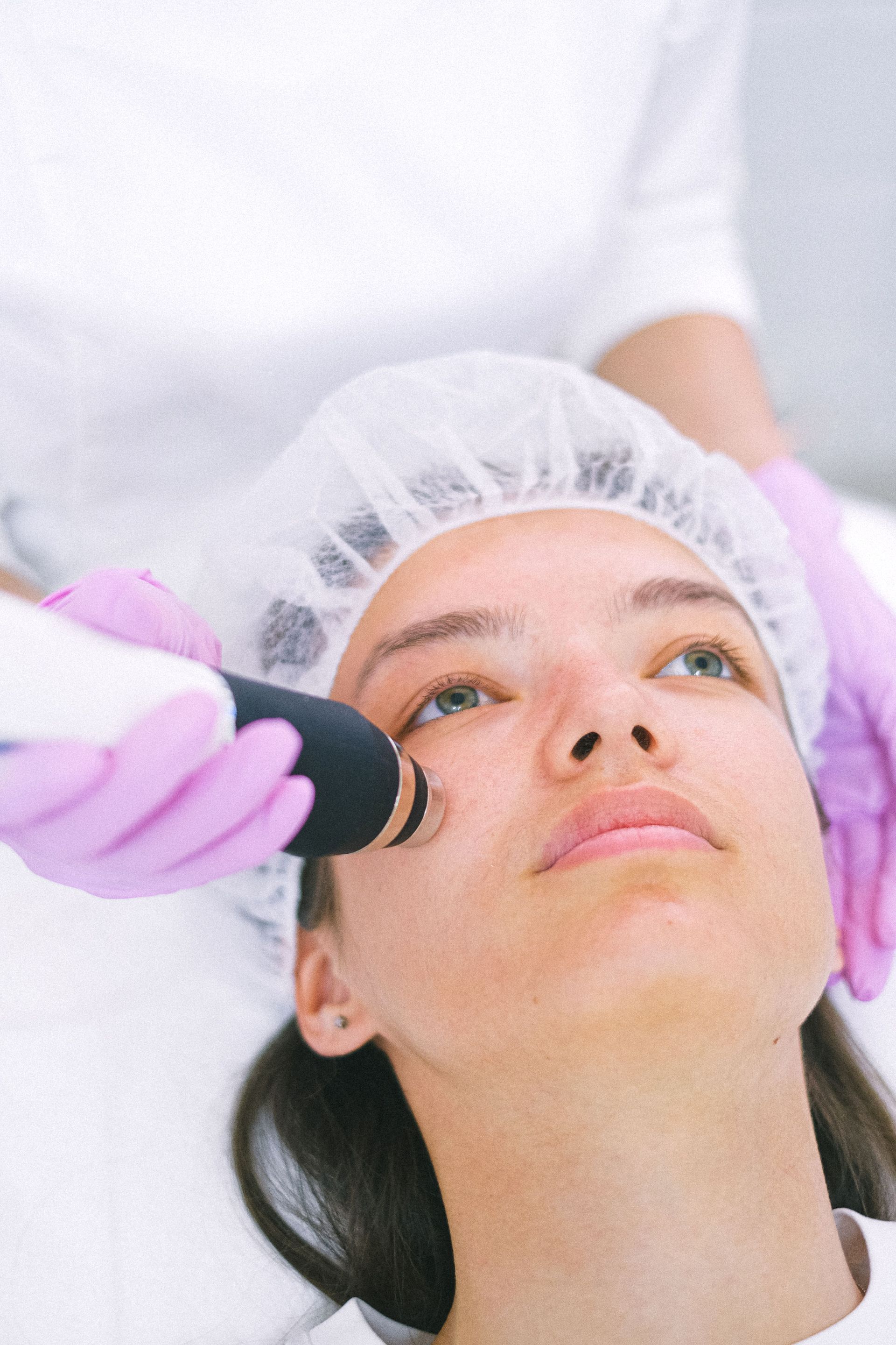 Facial treatment with gloved hands using a handheld device on a woman in a hair cap.