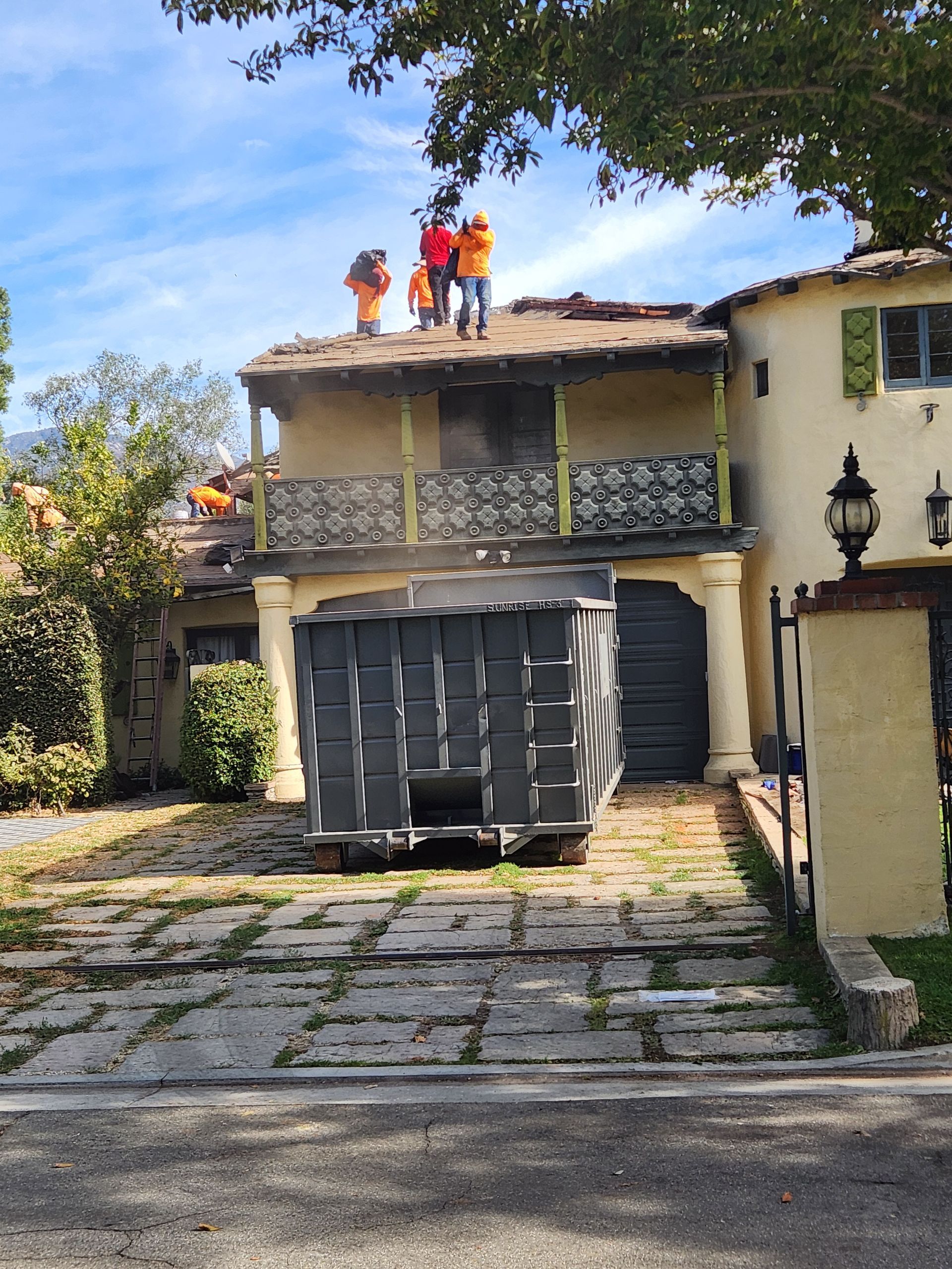 Roofers in orange vests work on a house roof next to a dumpster; sunny day.