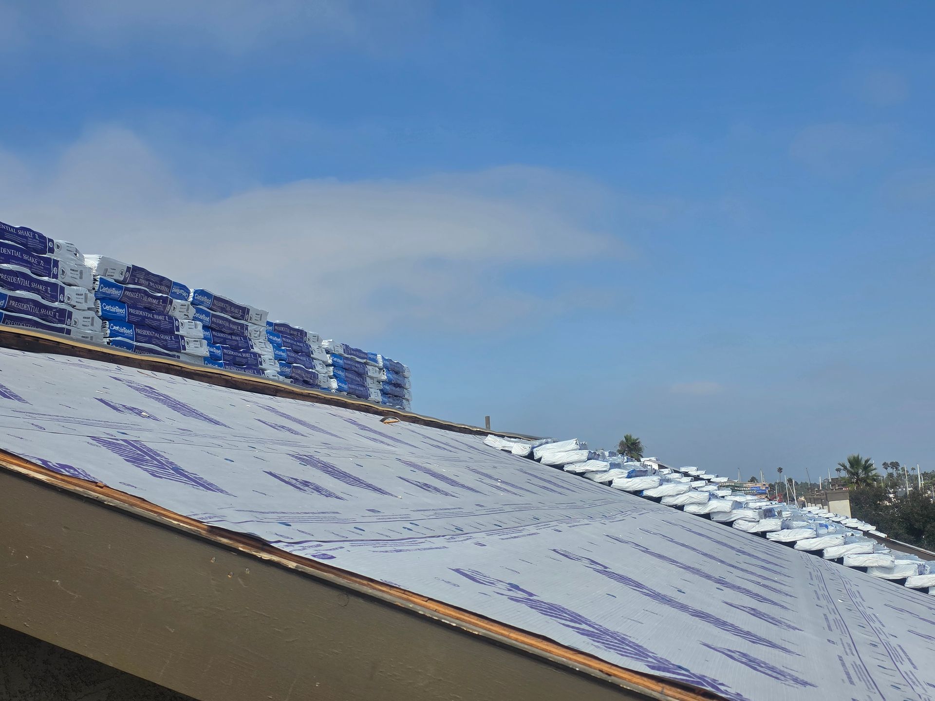 Rooftop with stacks of blue-wrapped shingles. Partial view of a bright, sunny sky above.