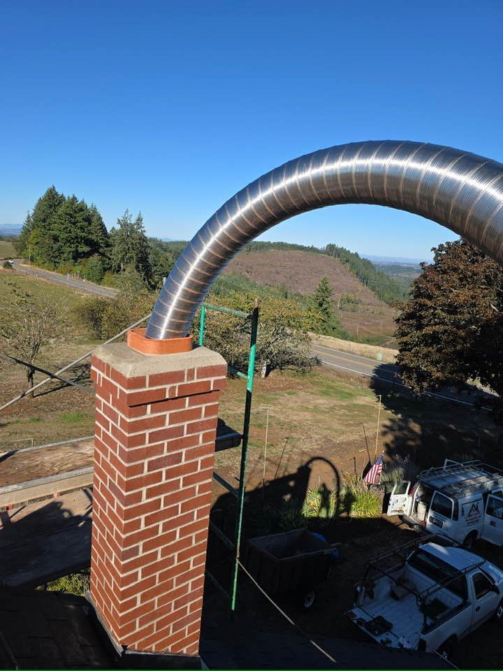 A flexible metal flue liner is being installed into a brick chimney against a backdrop of trees and a bright blue sky.