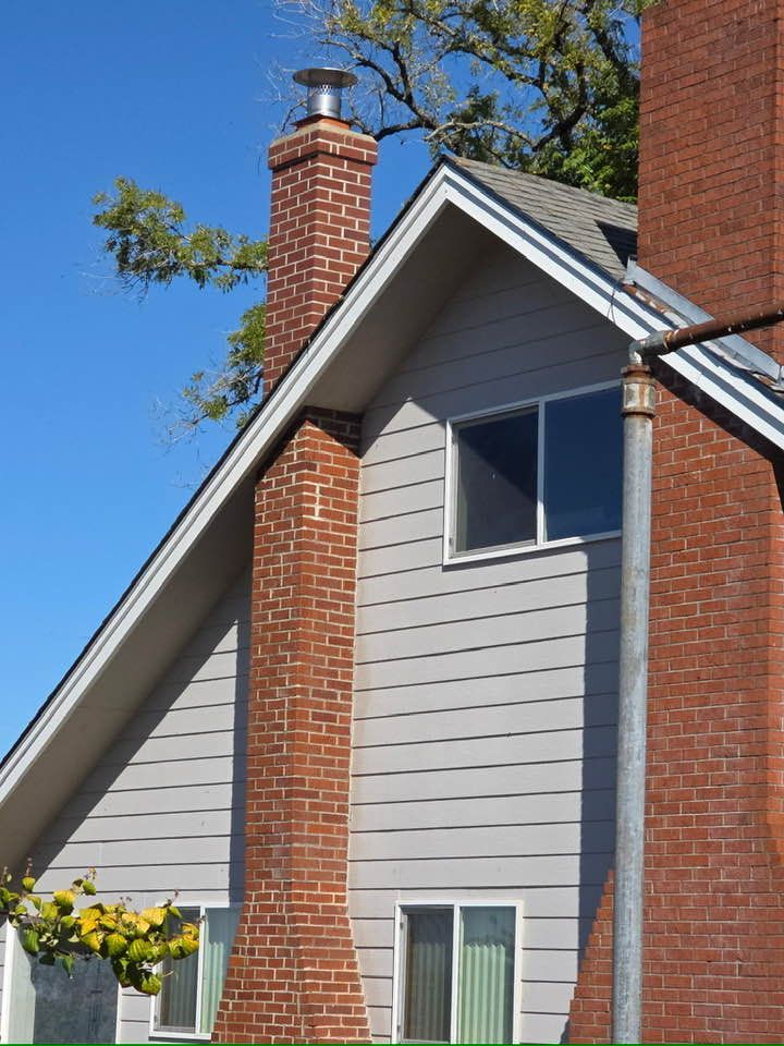 A gray-sided house with a steep gable roof and a red brick chimney, under a clear blue sky.