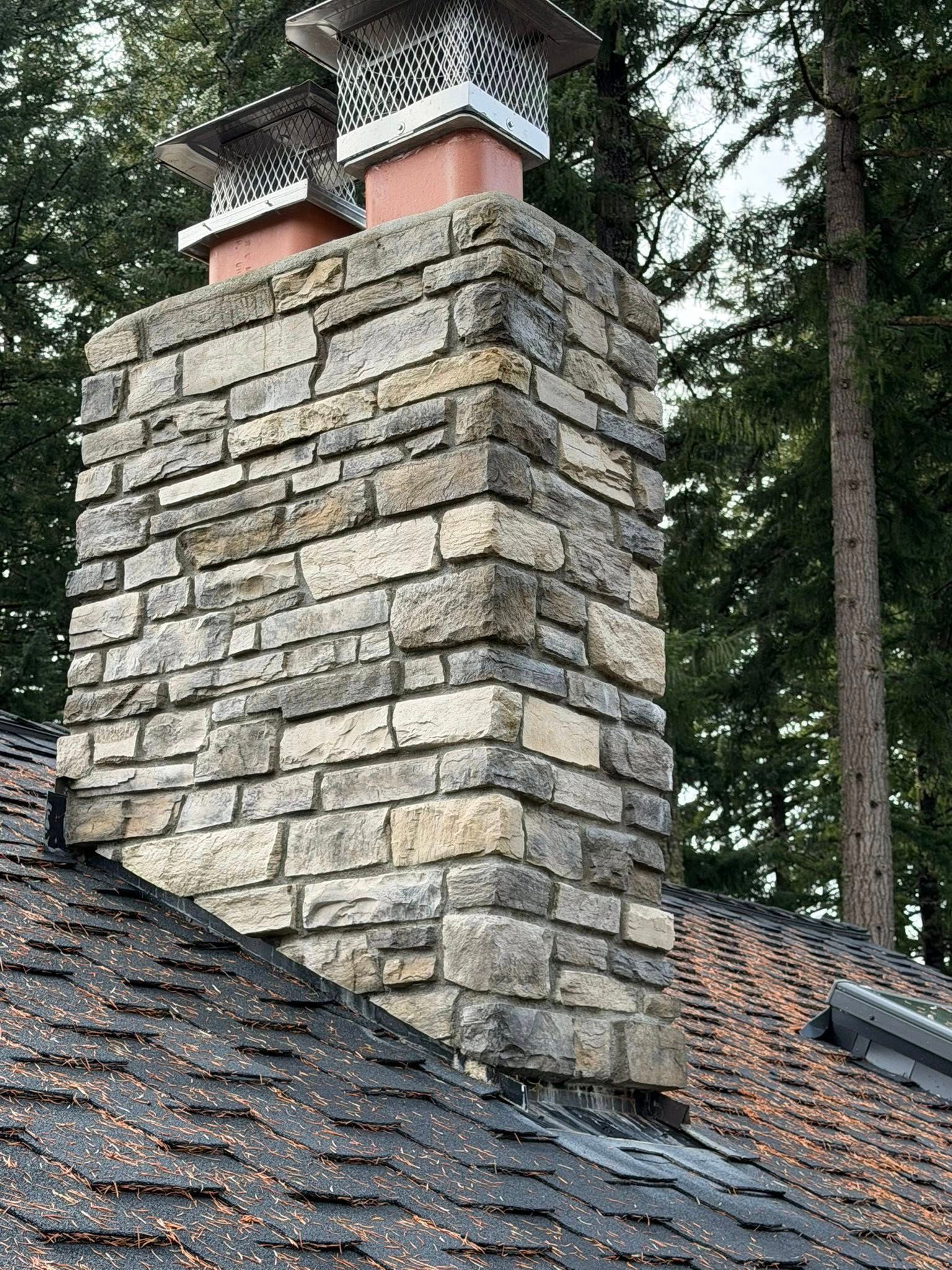 A stone chimney on a shingled roof with two metal chimney caps, set against a background of evergreen trees.