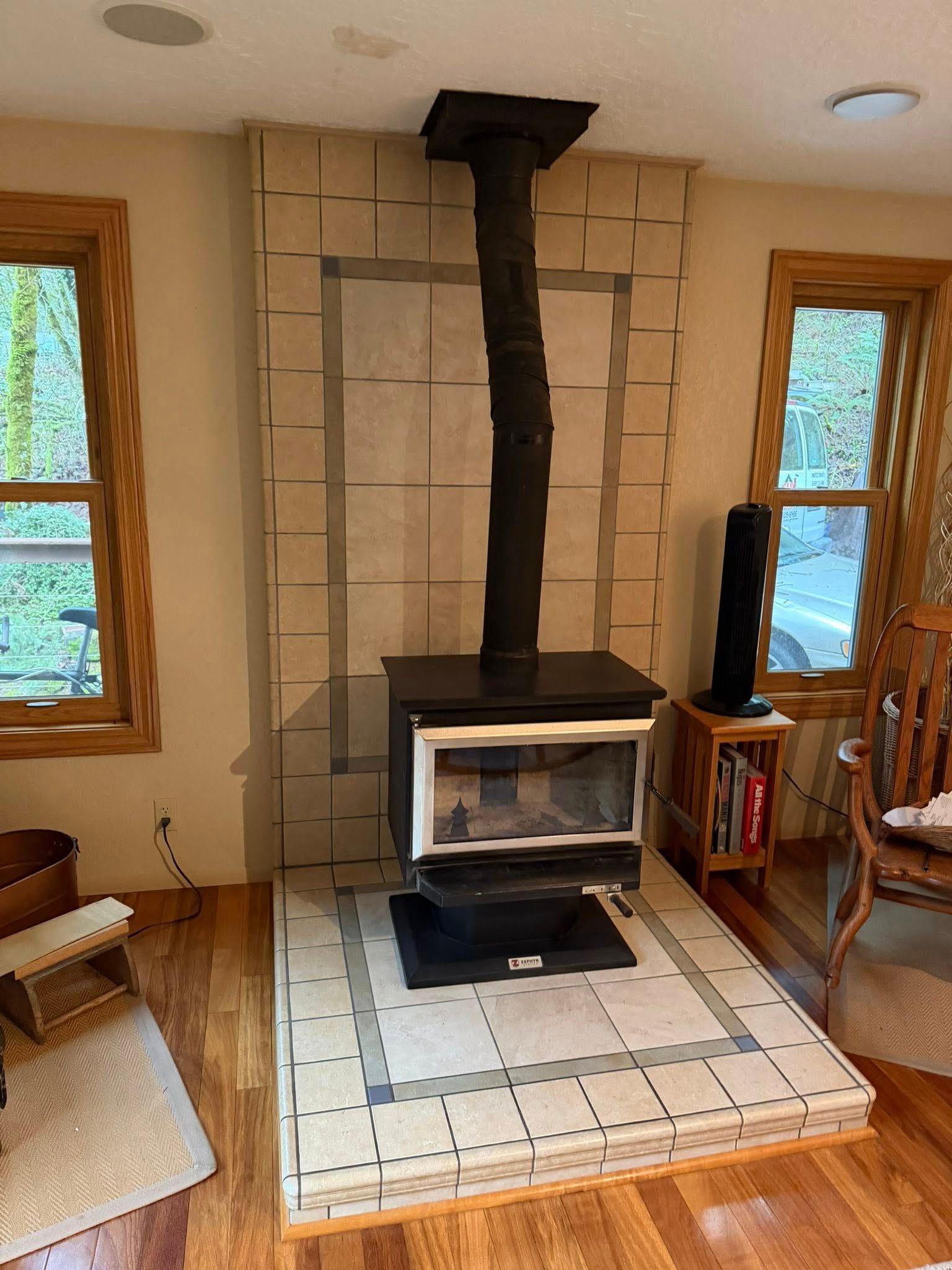 A wood-burning stove sits on a tiled hearth against a tall, light-colored tiled wall with a dark stovepipe flue.