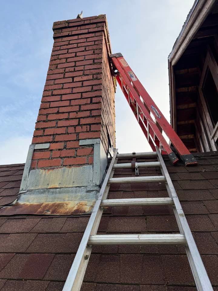 An aluminum extension ladder on a shingled roof leans against a brick chimney next to a red ladder.