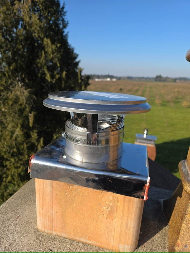A metal chimney cap sits atop a brick flue on a roof, with a blurred green landscape and blue sky in the background.