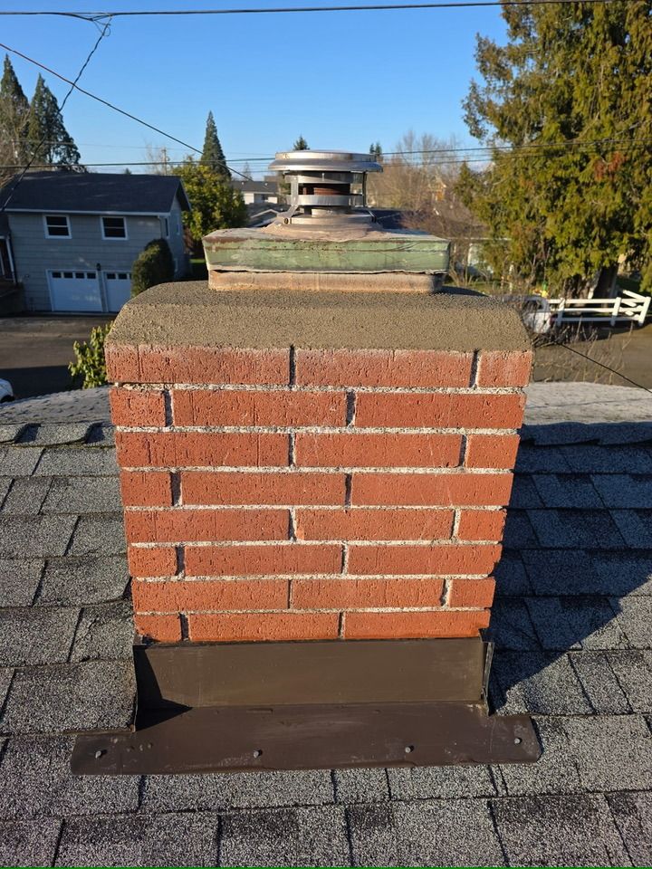 A red brick chimney with a metal cap and brown flashing, situated on a shingled roof under a clear blue sky.