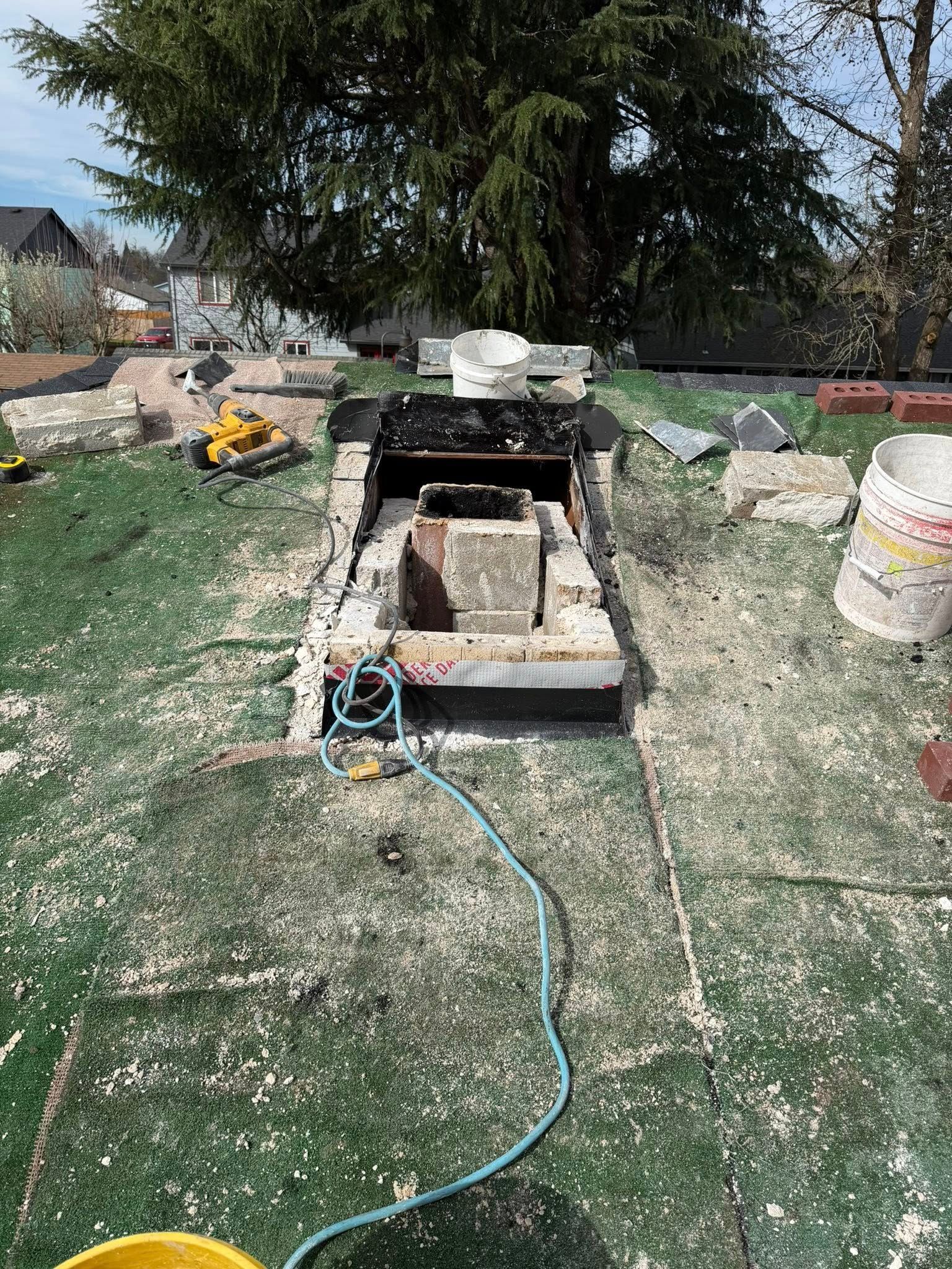 A brick chimney under repair on a rooftop covered in construction debris, with tools and a white bucket nearby.