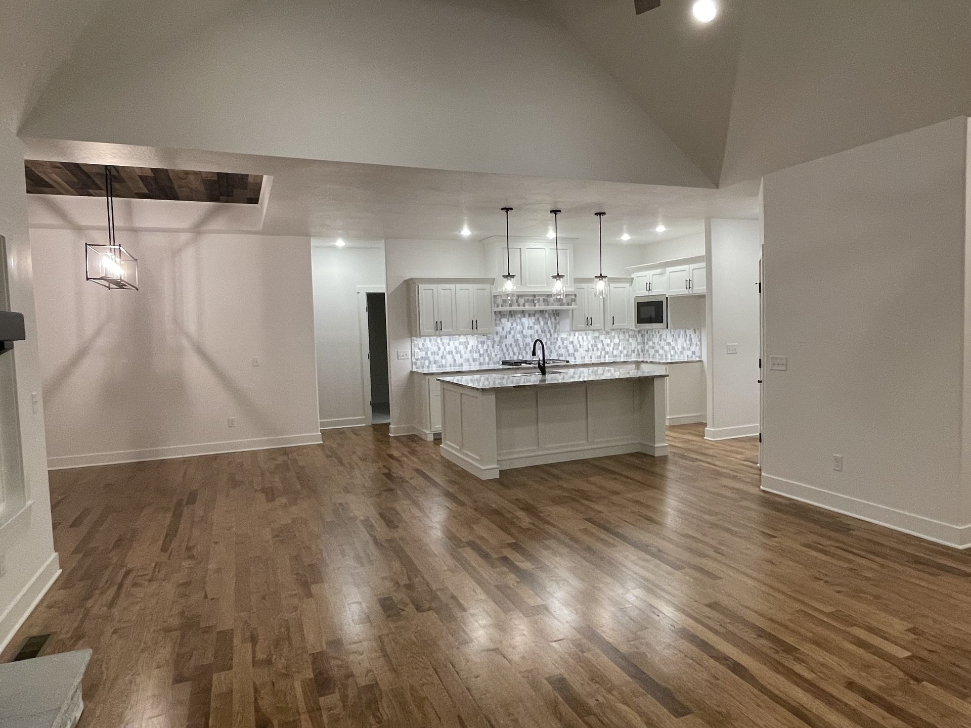 A large empty room with hardwood floors and a kitchen in the background.