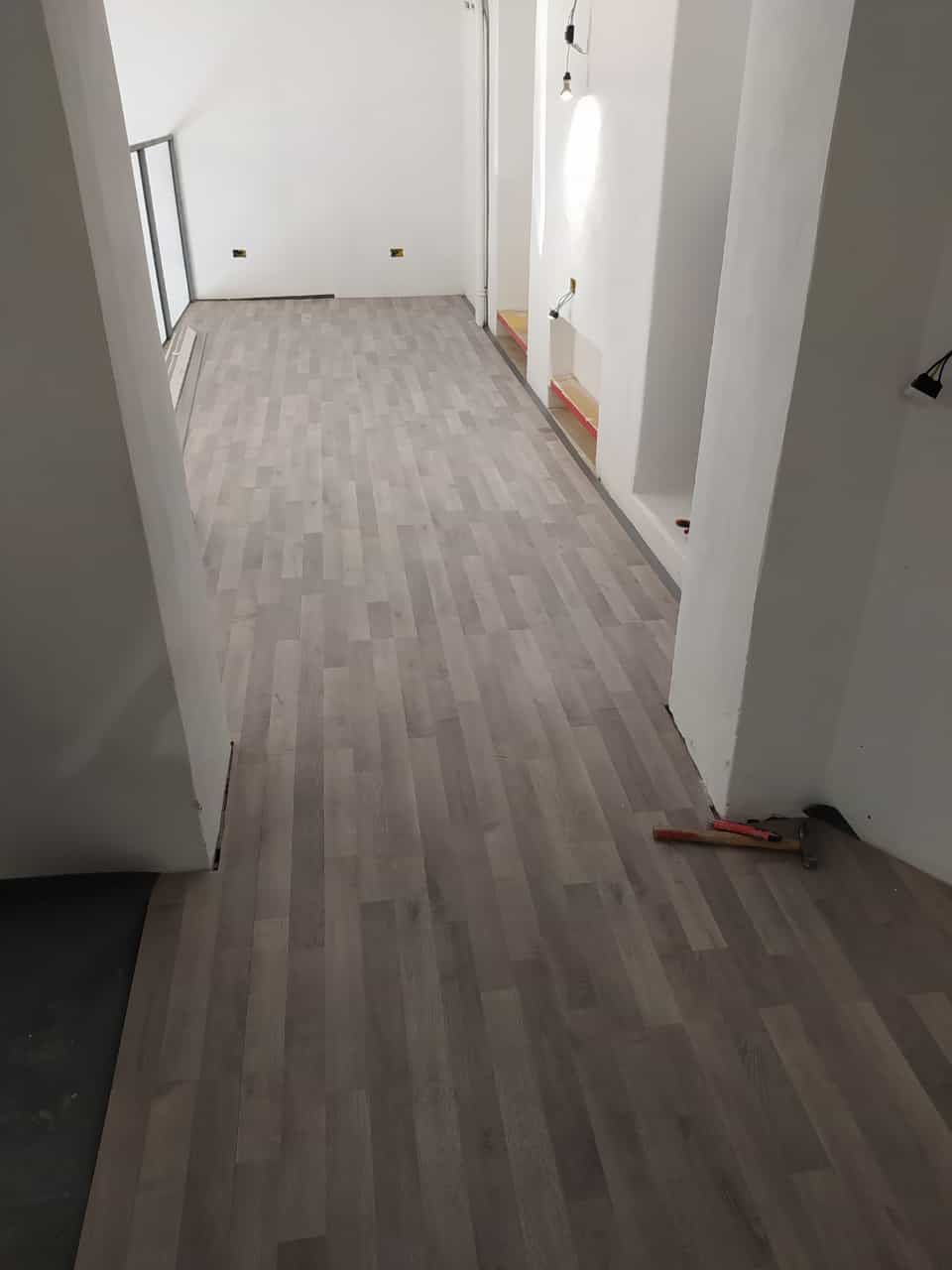 Stanza vuota con pavimento in legno grigio, pareti bianche e una piccola finestra. Attrezzi sparsi sul pavimento.