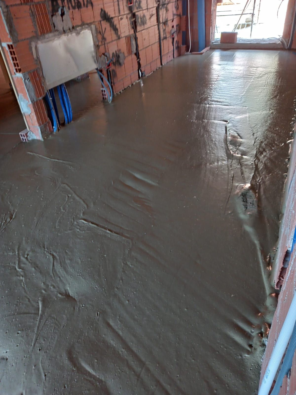 Pavimento in cemento appena gettato in una stanza in costruzione; sono visibili tubi blu e pareti in mattoni rossi.