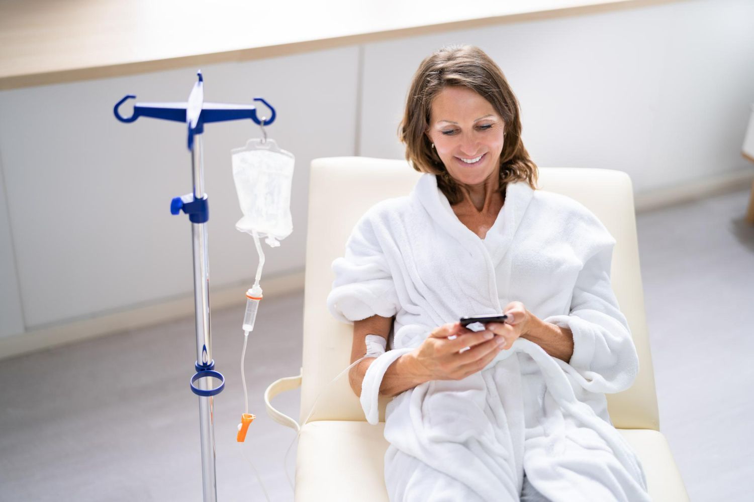 Woman in robe receiving IV treatment, smiling at phone in bright room.
