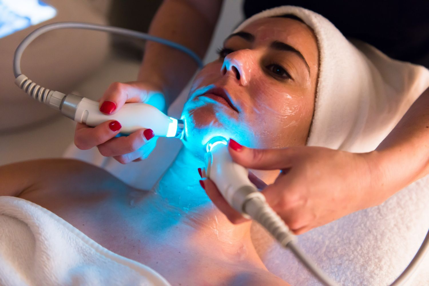 Woman receiving blue light therapy on her face in a spa setting, attended by a person holding the device.