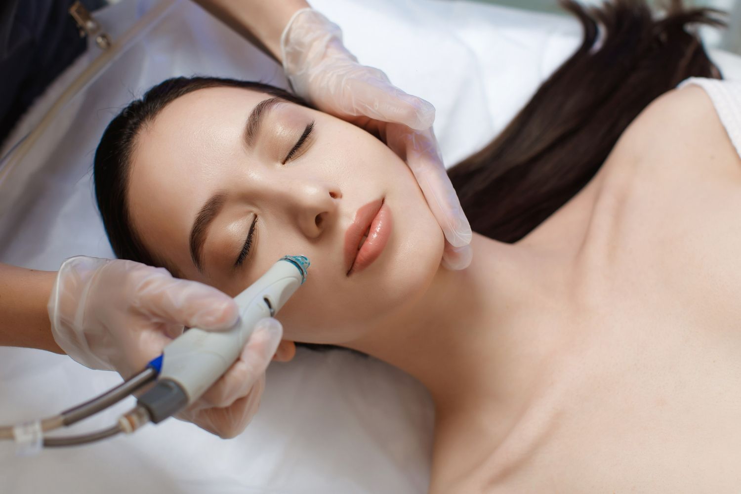 Woman receiving facial treatment, hands holding skin and device. Closed eyes, white sheet setting.