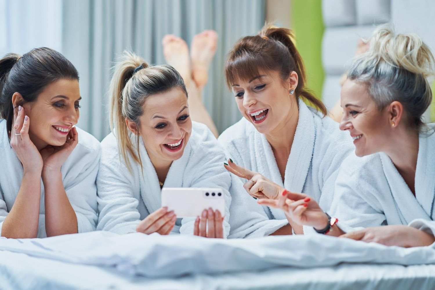 Four women in white robes laugh while looking at a phone on a bed.