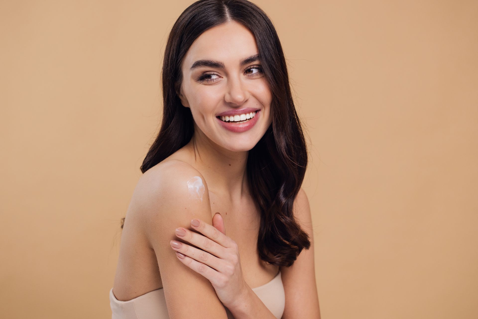 A woman is smiling and applying lotion to her shoulder.