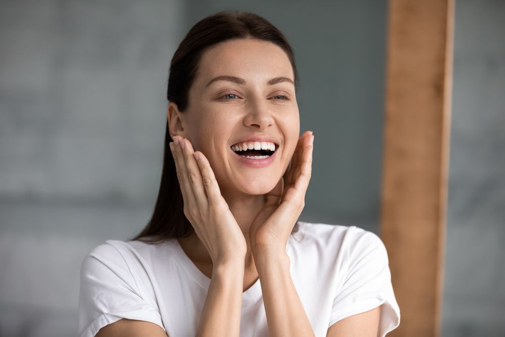 Woman in white t-shirt, smiling widely, hands on cheeks. Grey and wood background.