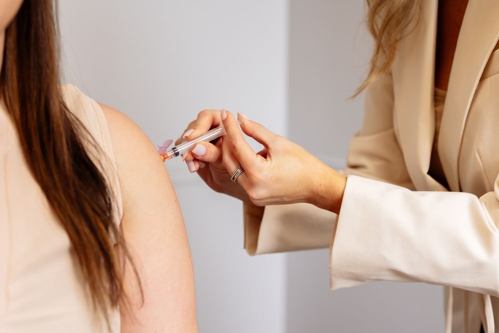 A person receives an injection in their upper arm by a person in a tan blazer.