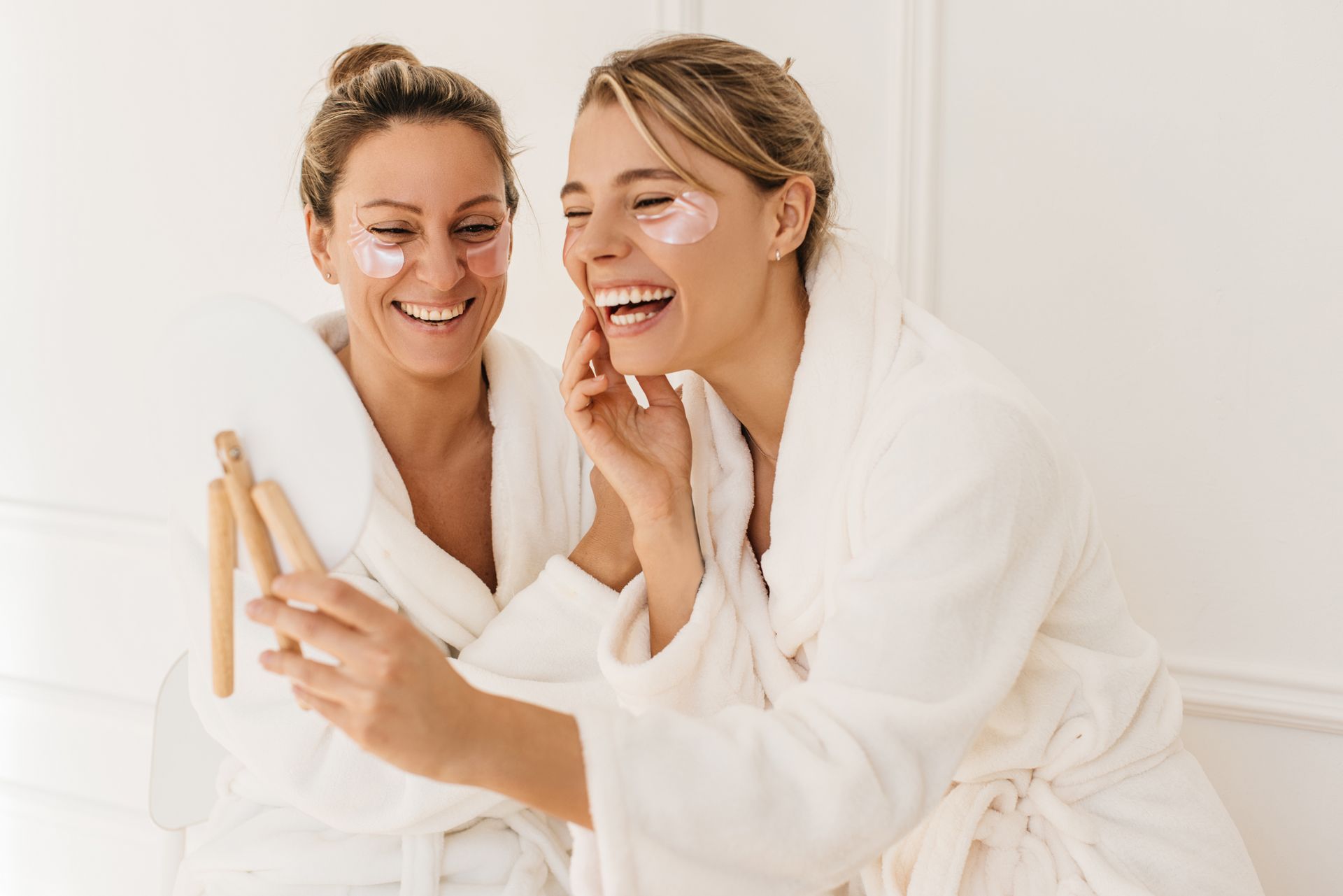 Two women in bathrobes are applying eye patches to their faces.