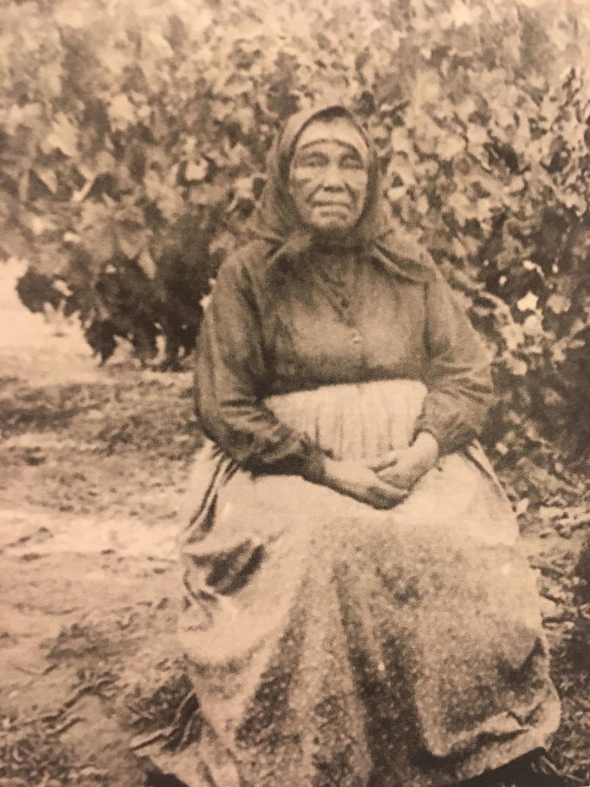 A black and white photo of an elderly woman sitting in front of a tree.