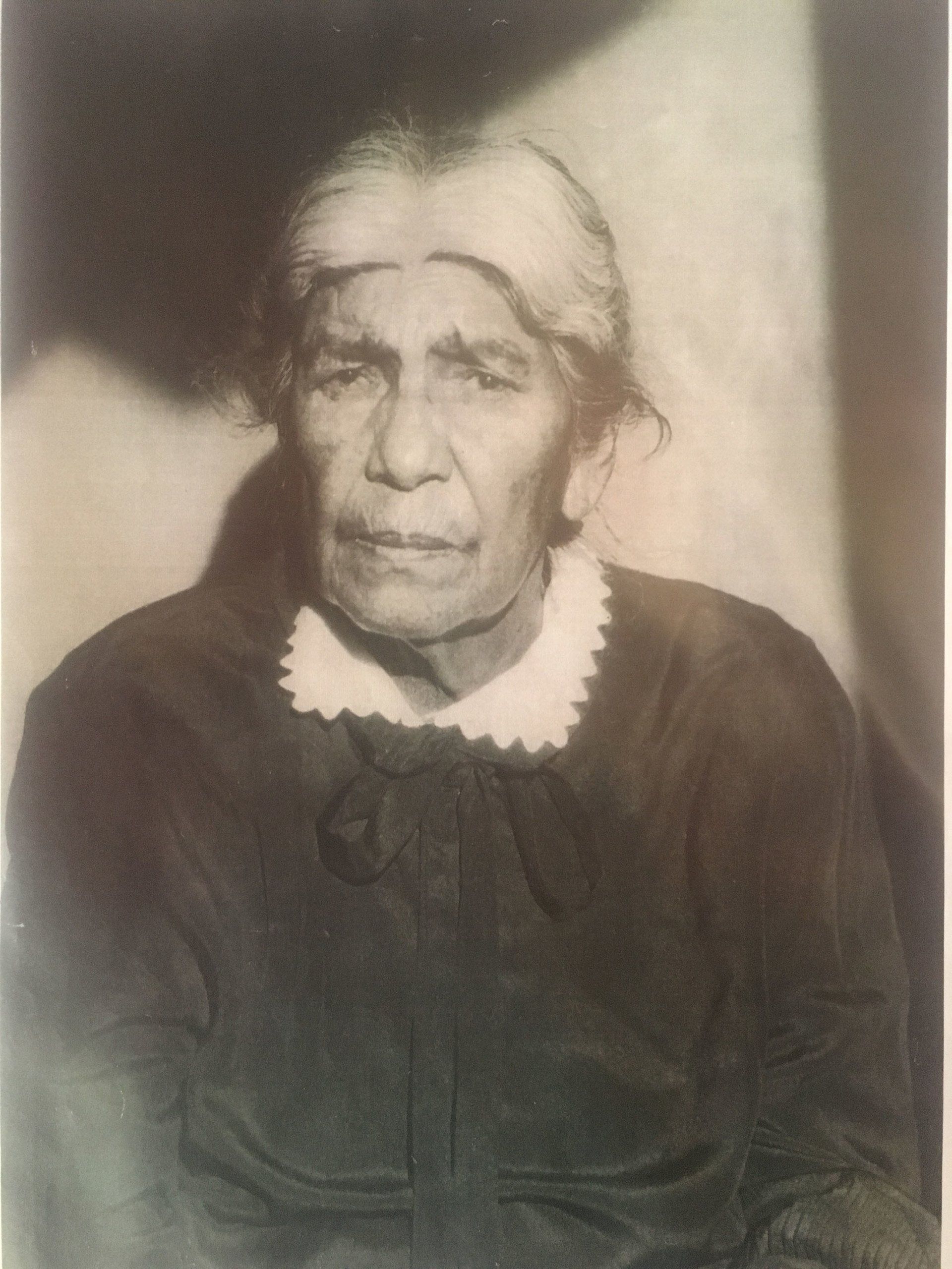 A black and white photo of an older woman wearing a black jacket and a white collar.
