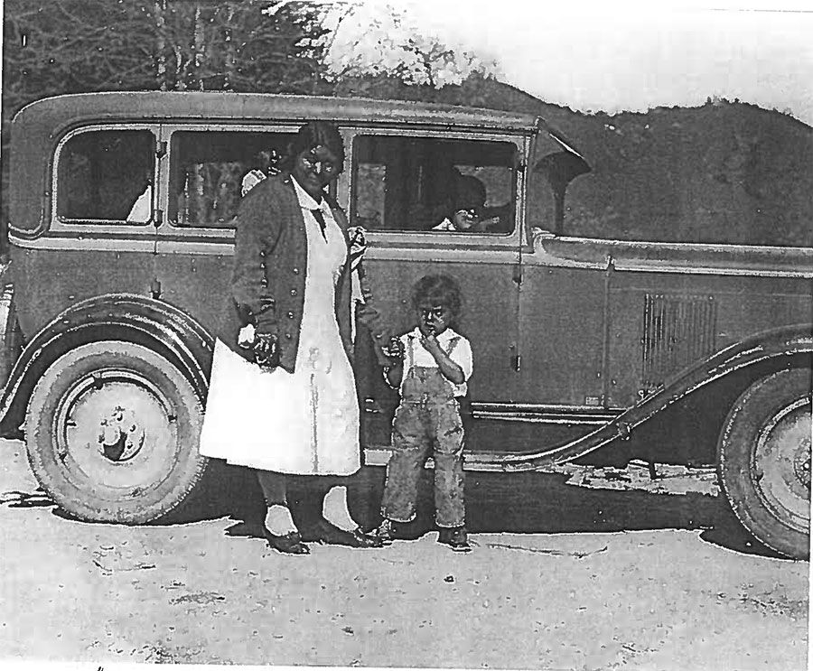 A black and white photo of a woman and child standing next to a car