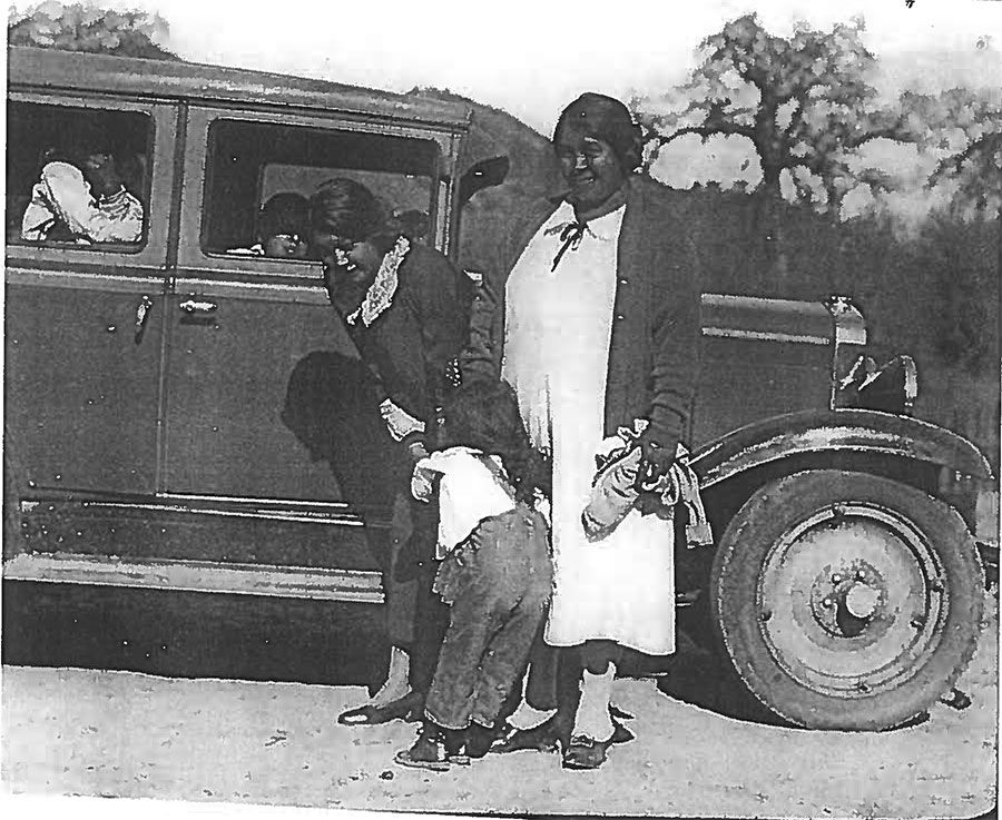 A black and white photo of a family standing next to a car