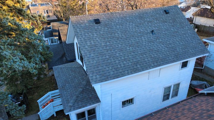 Aerial view of a white two-story house with a grey shingled roof, surrounded by trees and neighboring buildings.