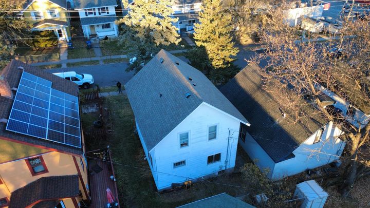 Aerial view of three houses, one with rooftop solar panels, surrounded by trees and a street on a sunny day.