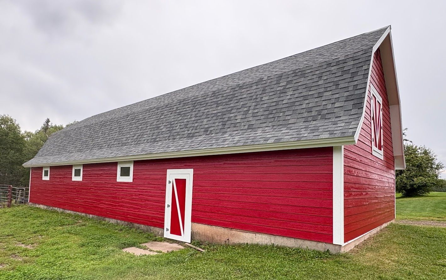 A long red barn with a grey shingled gambrel roof sits on a grassy field under a cloudy sky.